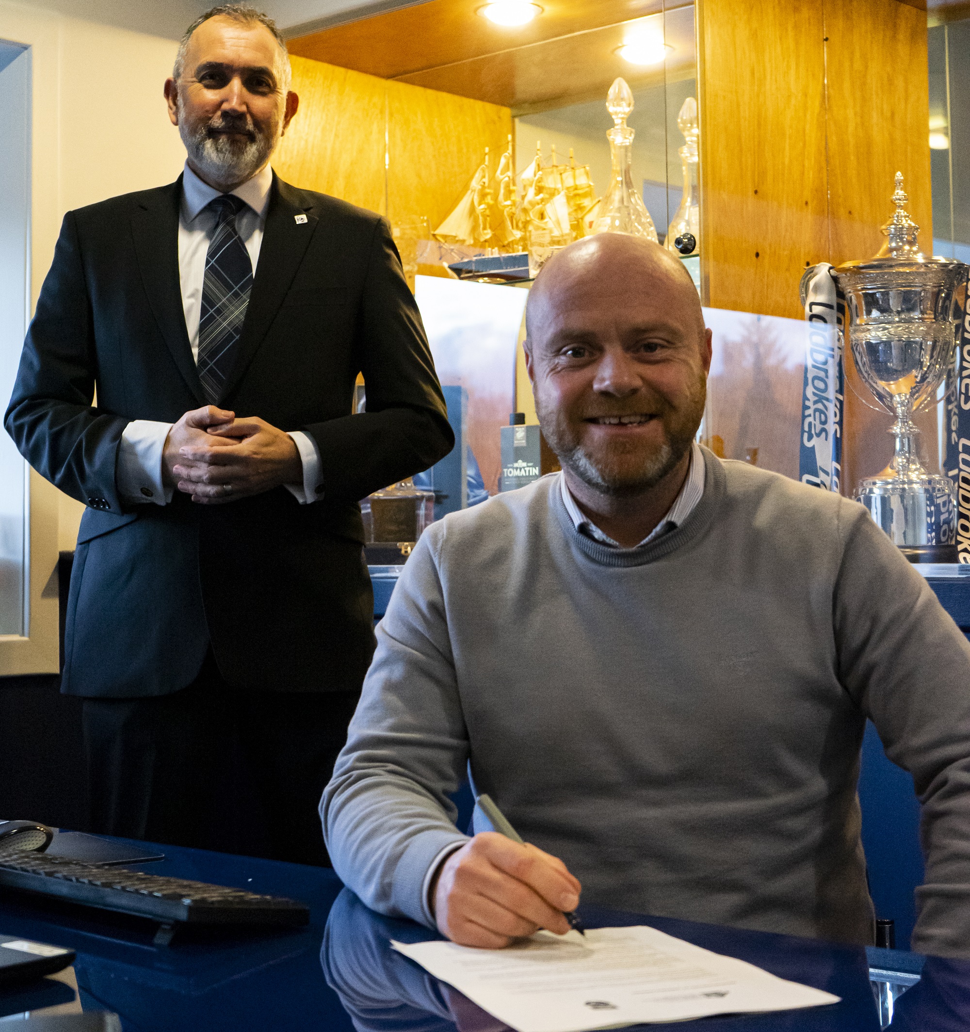 One person sitting with a pen signing a piece of paper. One person standing behind