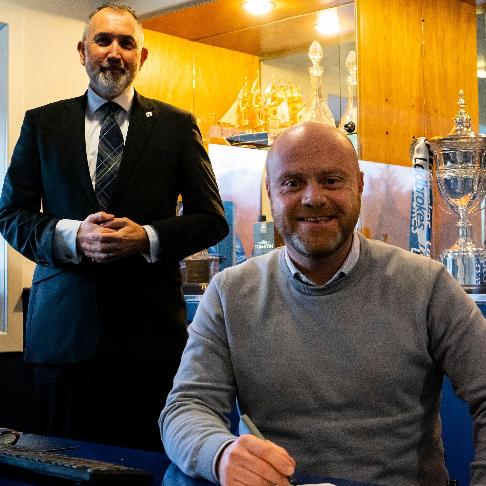 One person sitting with a pen signing a piece of paper. One person standing behind