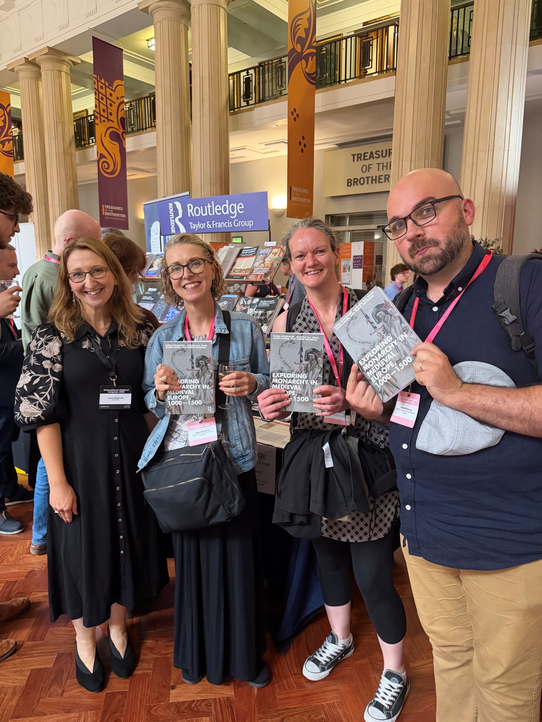 Four people at a publishing event, holding copies of Exploring Monarchy in Medieval Europe, 1000-1500, with a Routledge display in the background