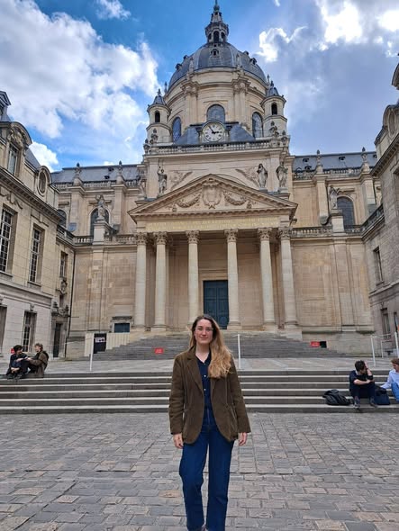 Woman standing on the steps leading towards a grand building with Corinthian columns supporting a decorated pediment, leading up towards a dome adorned with statues and a clockface.