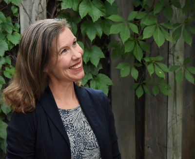 A smiling woman standing in the foreground of the image, with a fence with leafy vines behind her.