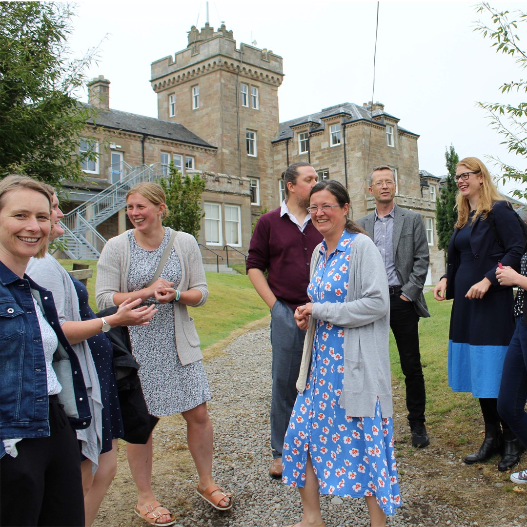 Group of people standing outside an old stone building, smiling and chatting.