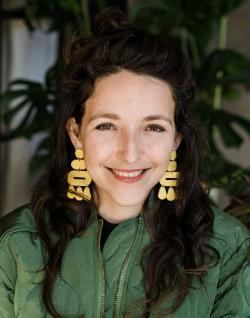 Close-up portrait photo of a woman smiling, wearing a green bomber-style jacket and large yellow earrings.