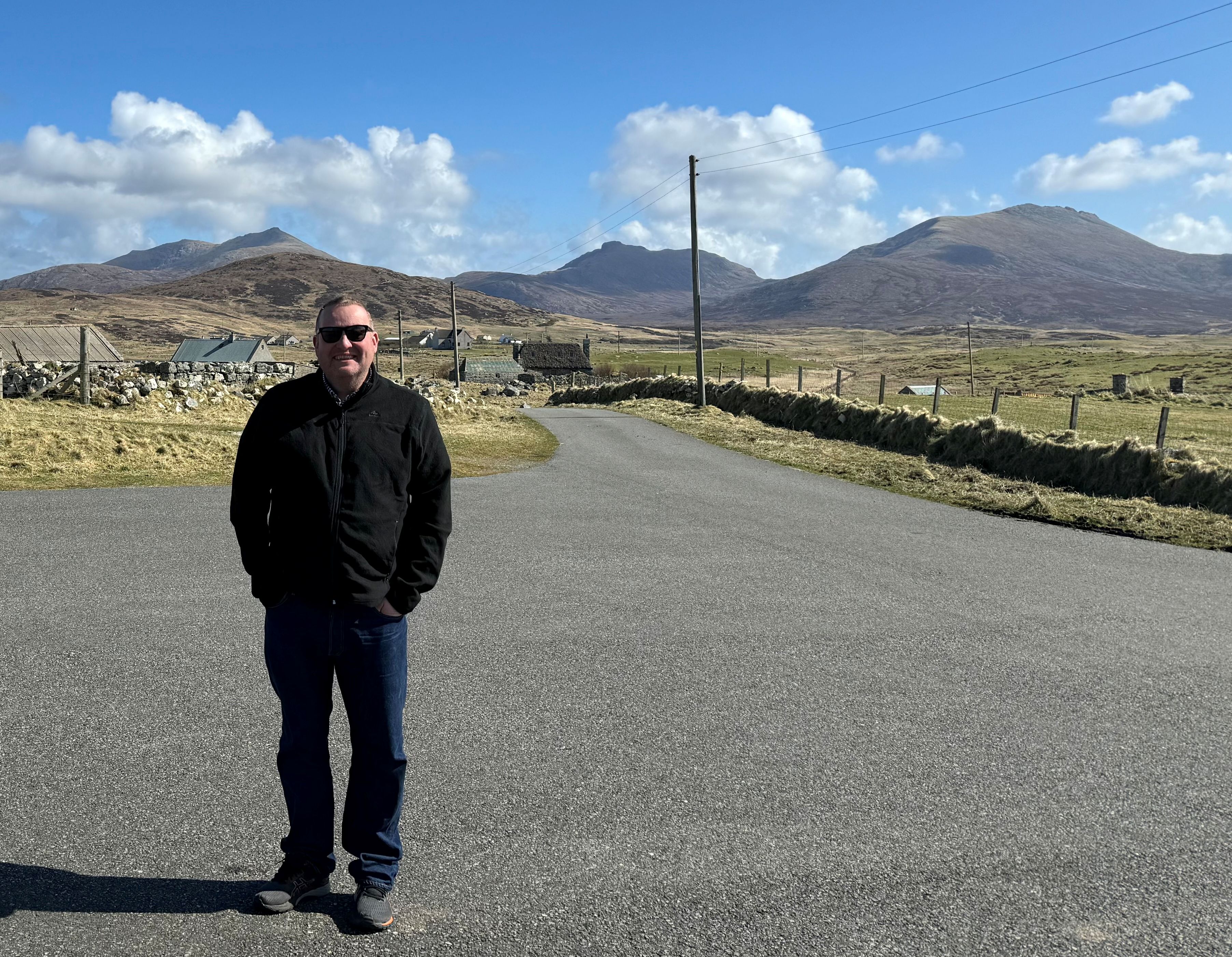 Person standing on a rural road with mountains and blue sky in the background.
